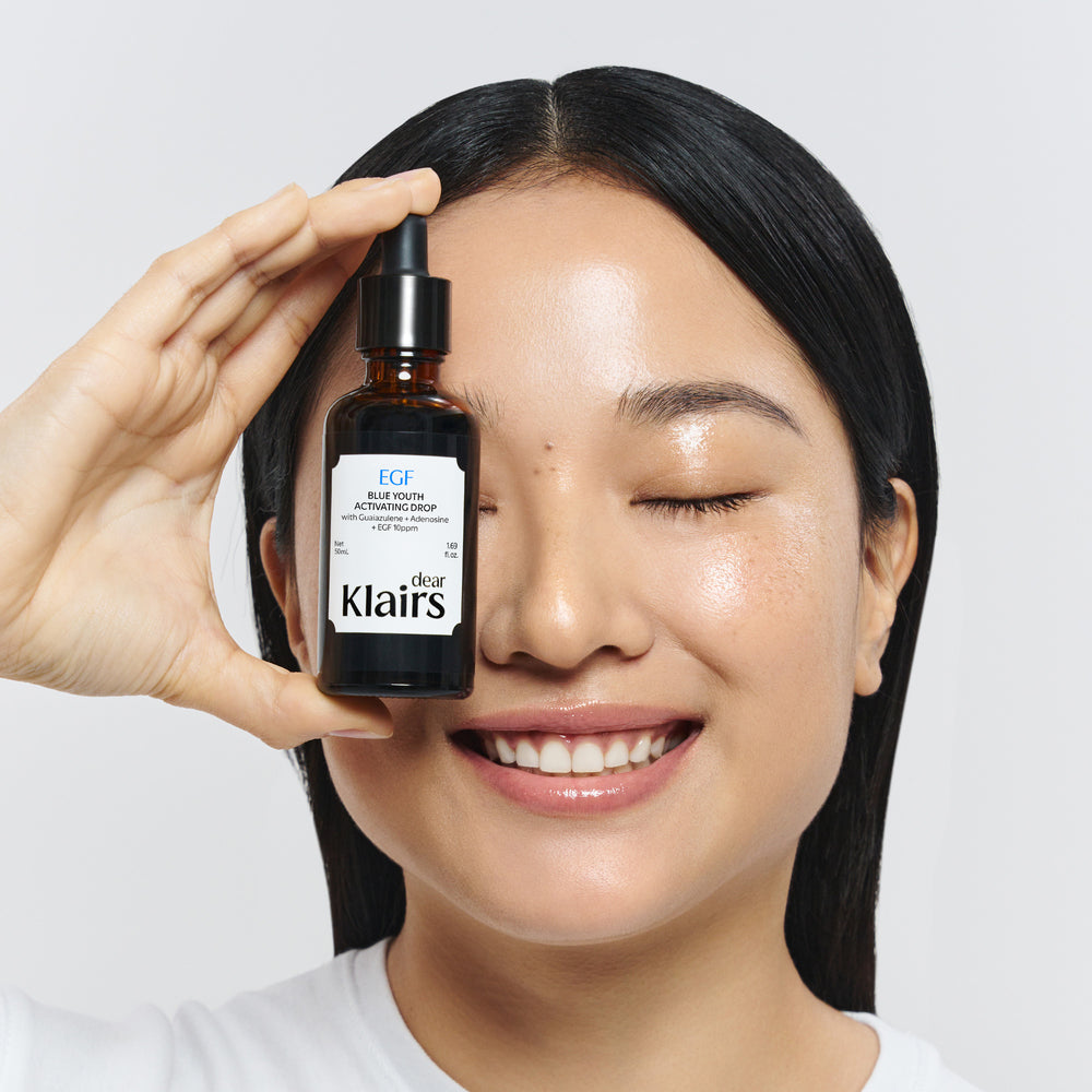 A woman with closed eyes holds a bottle of Dear Klairs EGF Blue Planet Repairing Serum in front of her face against a light gray background. Korean face serum for glowing skin, LUME