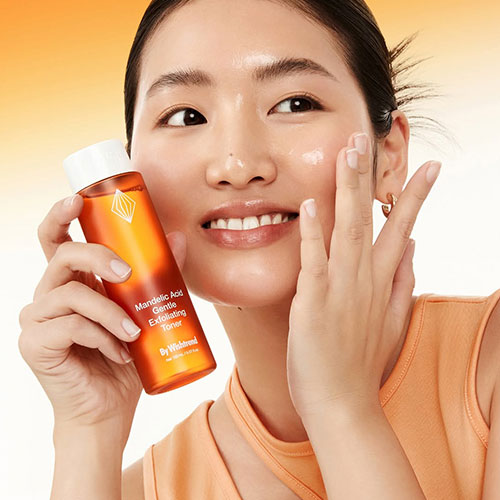 A woman with glowing skin holds a bottle of Bywishtrend Mandelic Acid Exfoliating Toner while this bright orange toner bottle to her cheek against a warm gradient background. Gentle exfoliator for smooth skin, LUME
