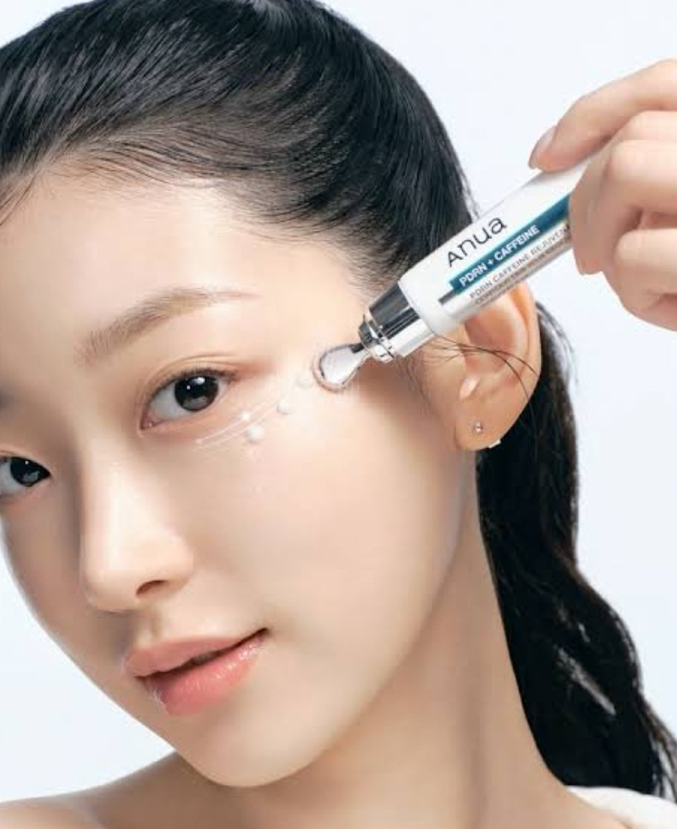 A close-up of a woman's face as she applies a gel cushion from an Anua Eye Careline skincare tube to her cheek, with her hand holding the applicator visible on the right. Korean eye care cream for the delicate eye area, LUME