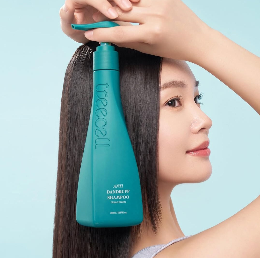 A woman with long, straight dark hair hold Treecell Anti dandruff shampoo from a teal bottle to her hair against a light blue background. Korean hair care products for healthy hair and scalp, LUME