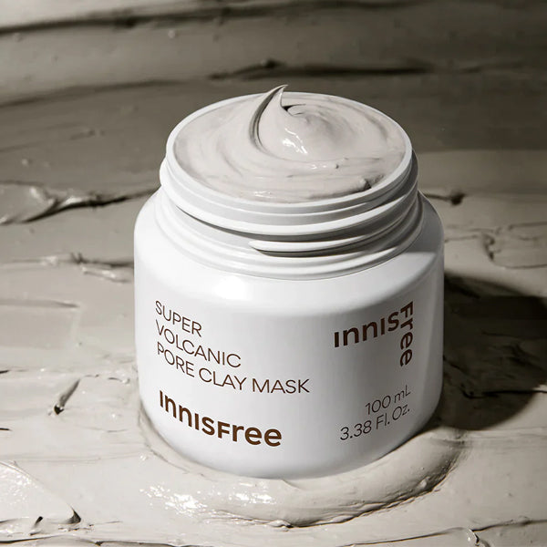 An open white jar of Innisfree Super Volcanic Pore Clay Mask, showing the thick gray clay mask cream swirled on top, set against a textured background of the same clay mask. Clay mask for deep pore cleansing, LUME